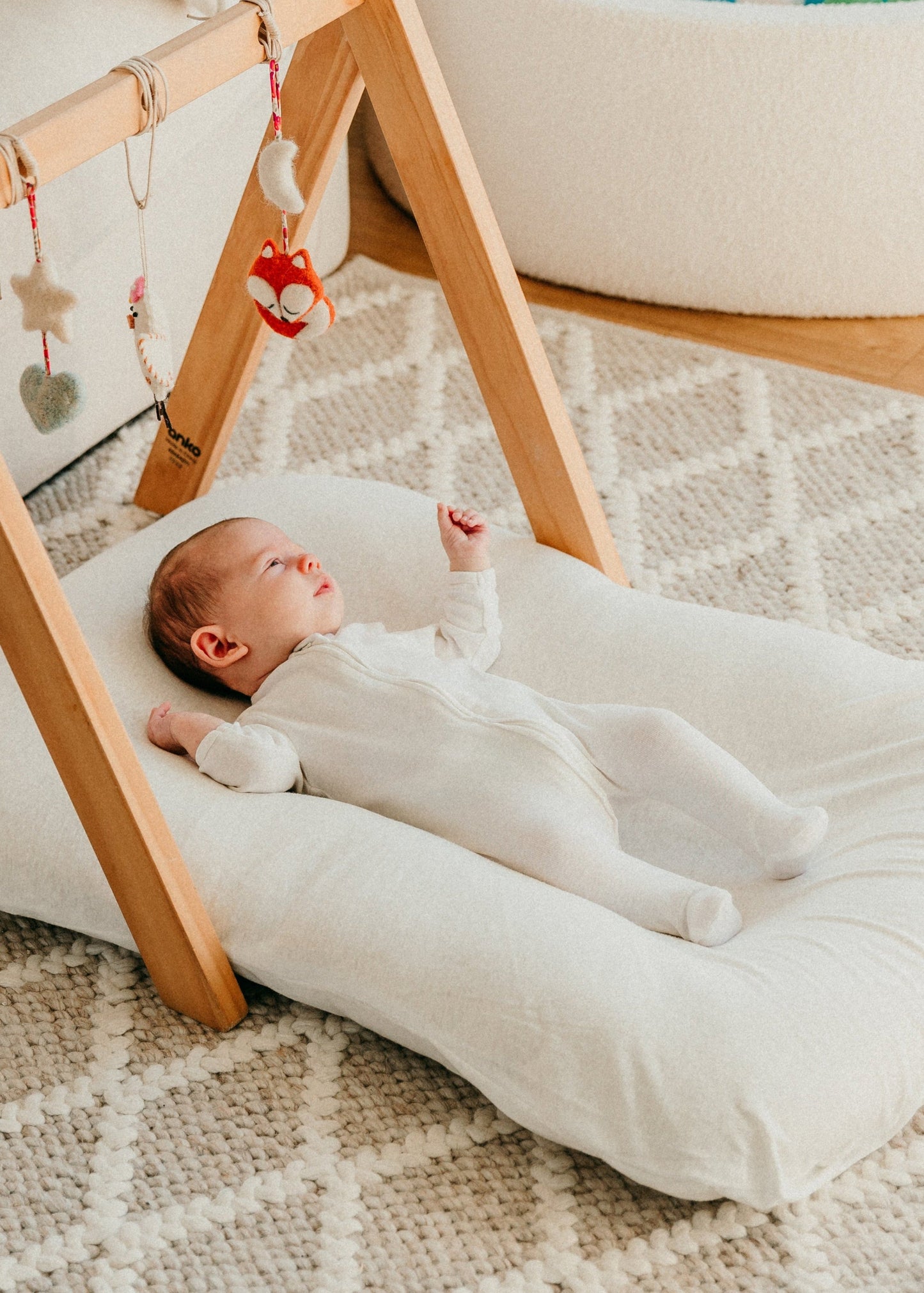 Baby lying on a white cushion next to a wooden play gym with colorful balls in the background.