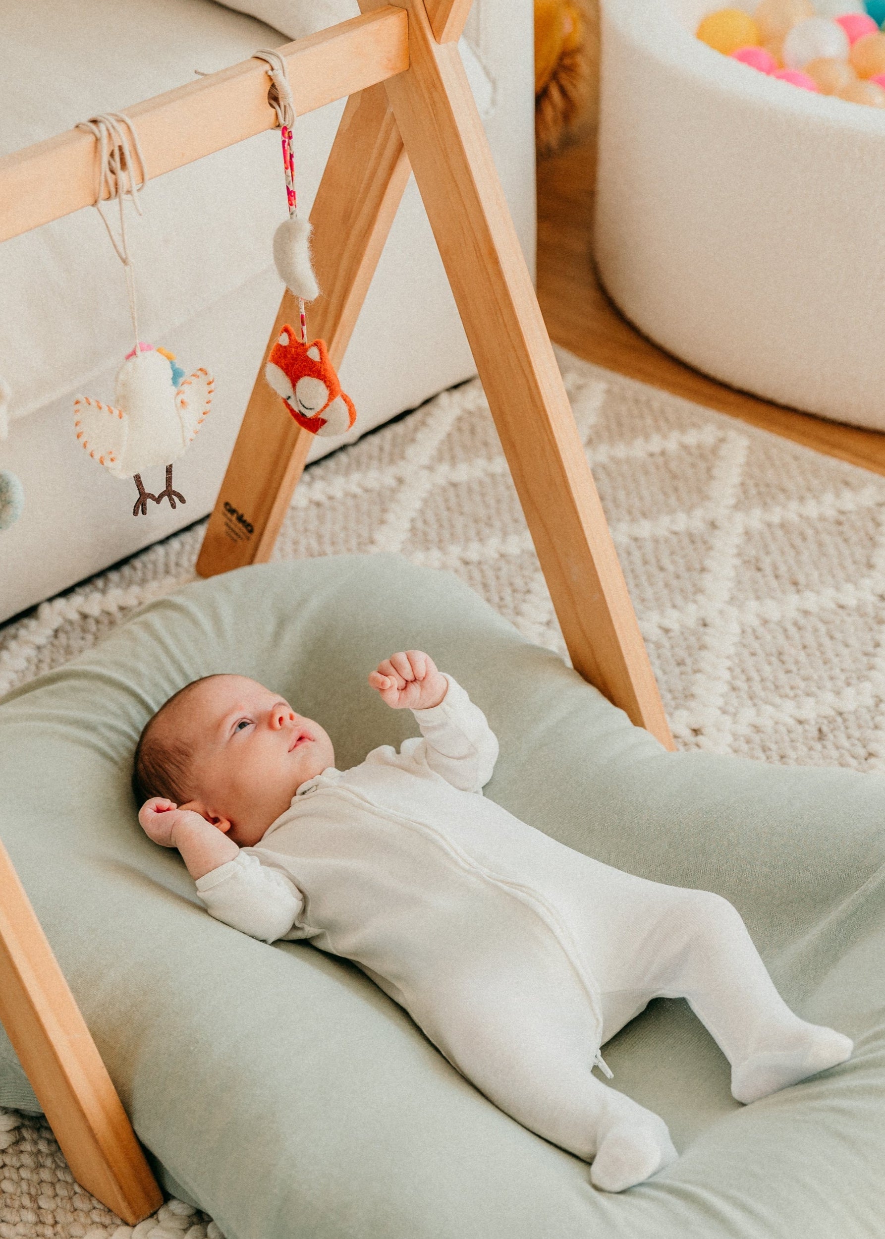 Baby lying on a green cushion next to a wooden baby gym with hanging toys in a room with a ball pit.